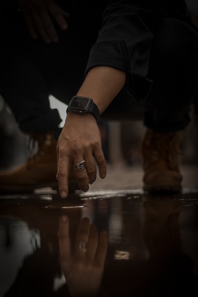 A person touches a water reflection while kneeling, wearing rings and a smartwatch.